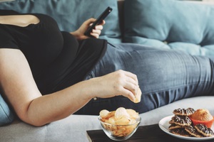 obese woman laying on sofa with smartphone eating chips learning the warning signs of diabetes