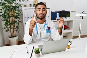A male doctor giving information about asthma from his laptop while holding an inhaler and a stethoscope