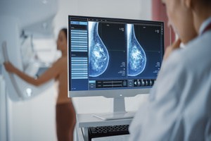 A doctor checking a live mammogram of breast tissues on a screen of a woman standing topless in the distance