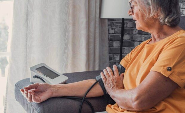 senior elder woman measure high low blood pressure test holding using medical digital electronic meter sit on sofa at home