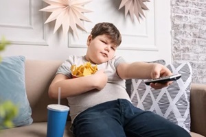 overweight boy watching TV with snacks indoors