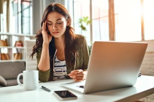 tired business woman in stress works at a laptop while sitting at a table at home and holds her hand on her temples