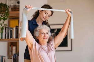 senior patient doing exercise with elastic band at home with personal trainer